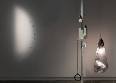 A sculpture with a hanging light stands upright in the room with nightshifts written as an arc on the wall behind.