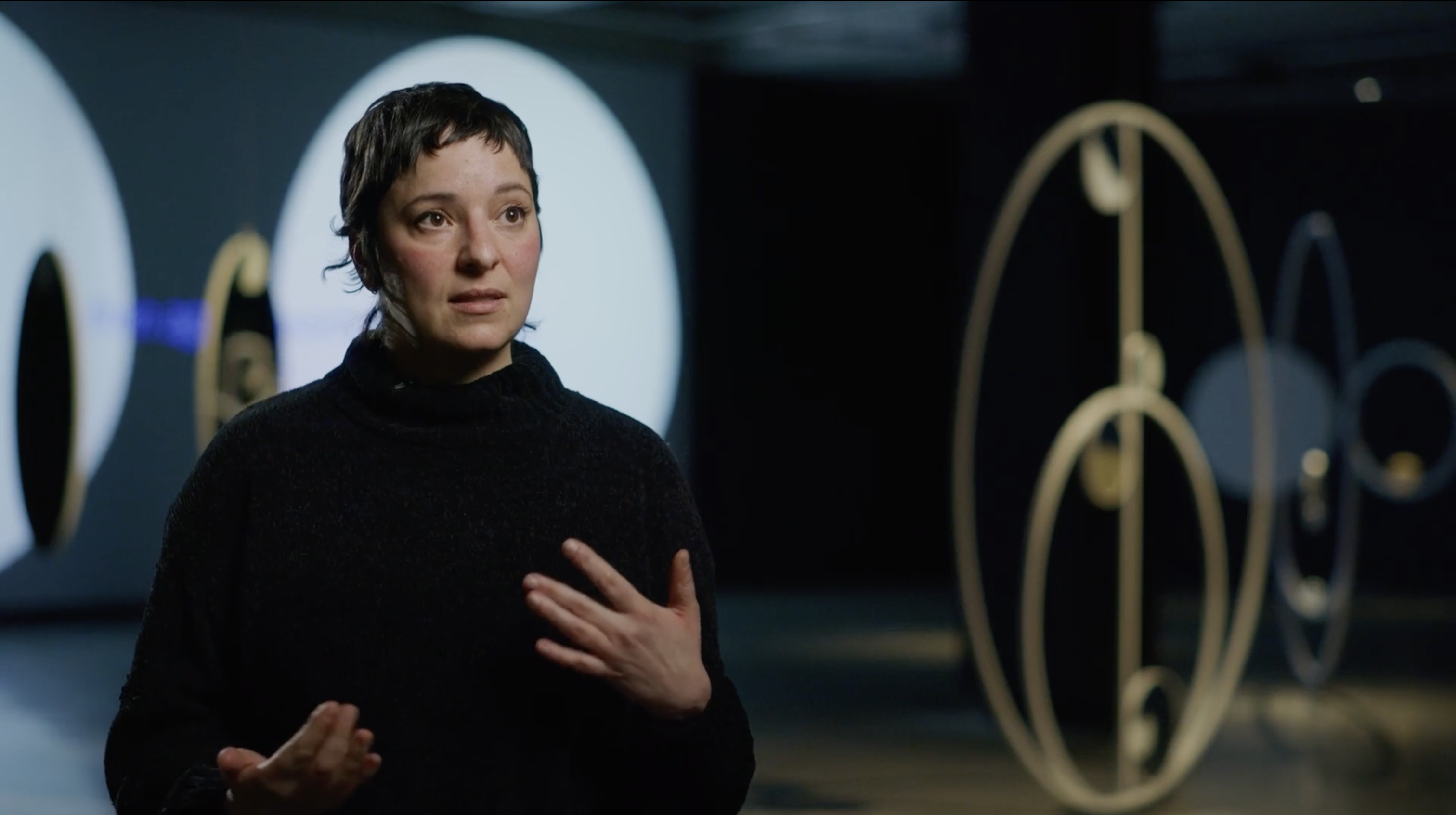 Artist Lisa Sammut stands in the gallery in front of her artwork as she speaks to an interviewer