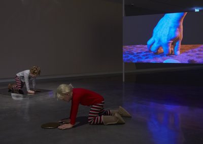 Sculptures of two children on hands and knees in a gallery with a video projection in background