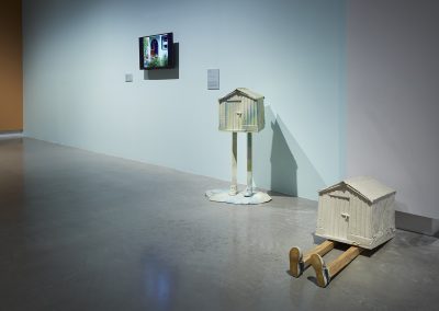 Two sculptures of small sheds with legs in a gallery
