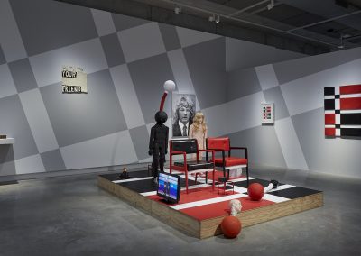 An exhibit of red and black chairs with a screen sits on a plinth in the middle of a gallery