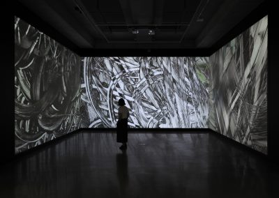 A person stands in front of a 3 channel projection in a dark room