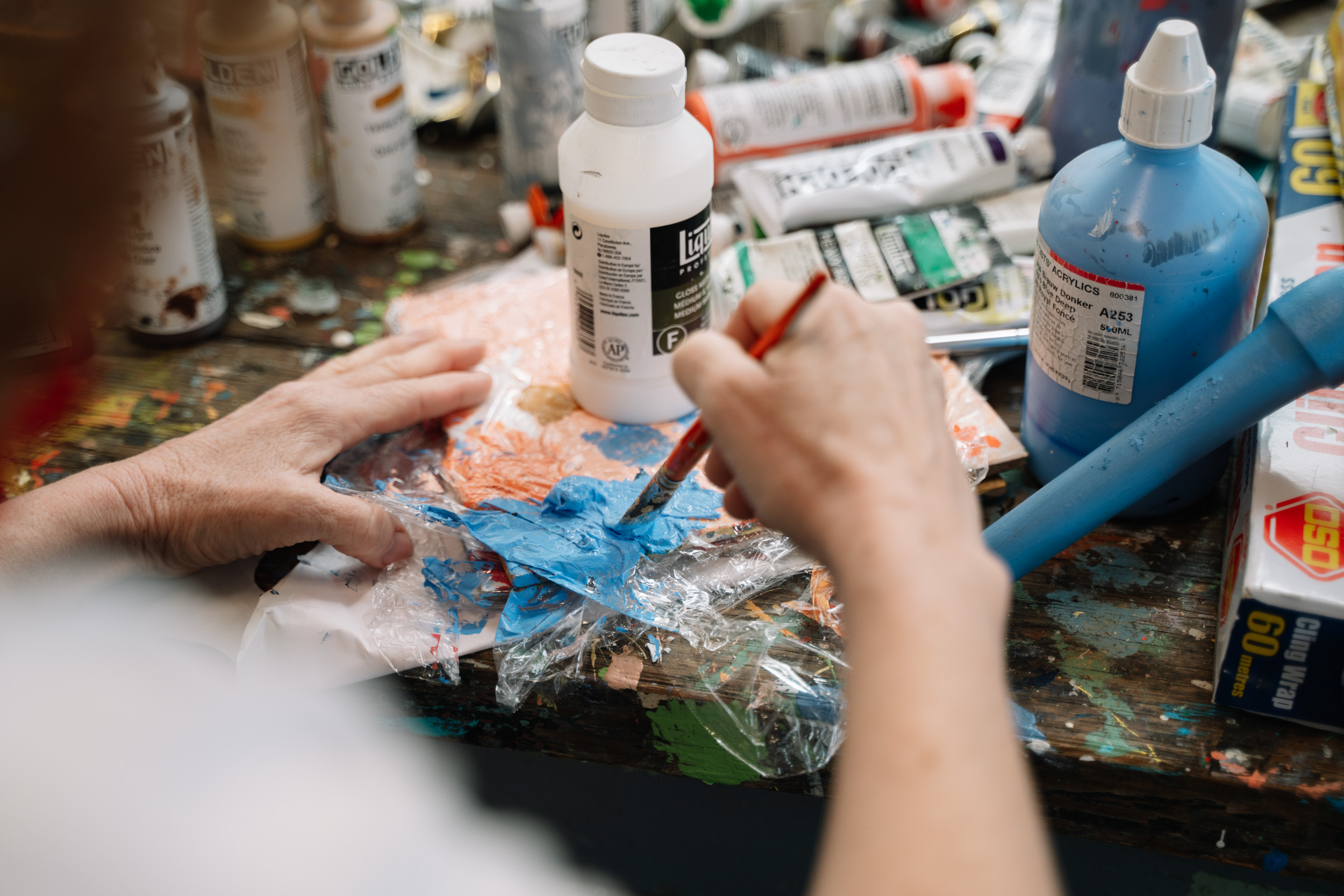 An artist mixes blue paint on glad wrap on a desk full of paints