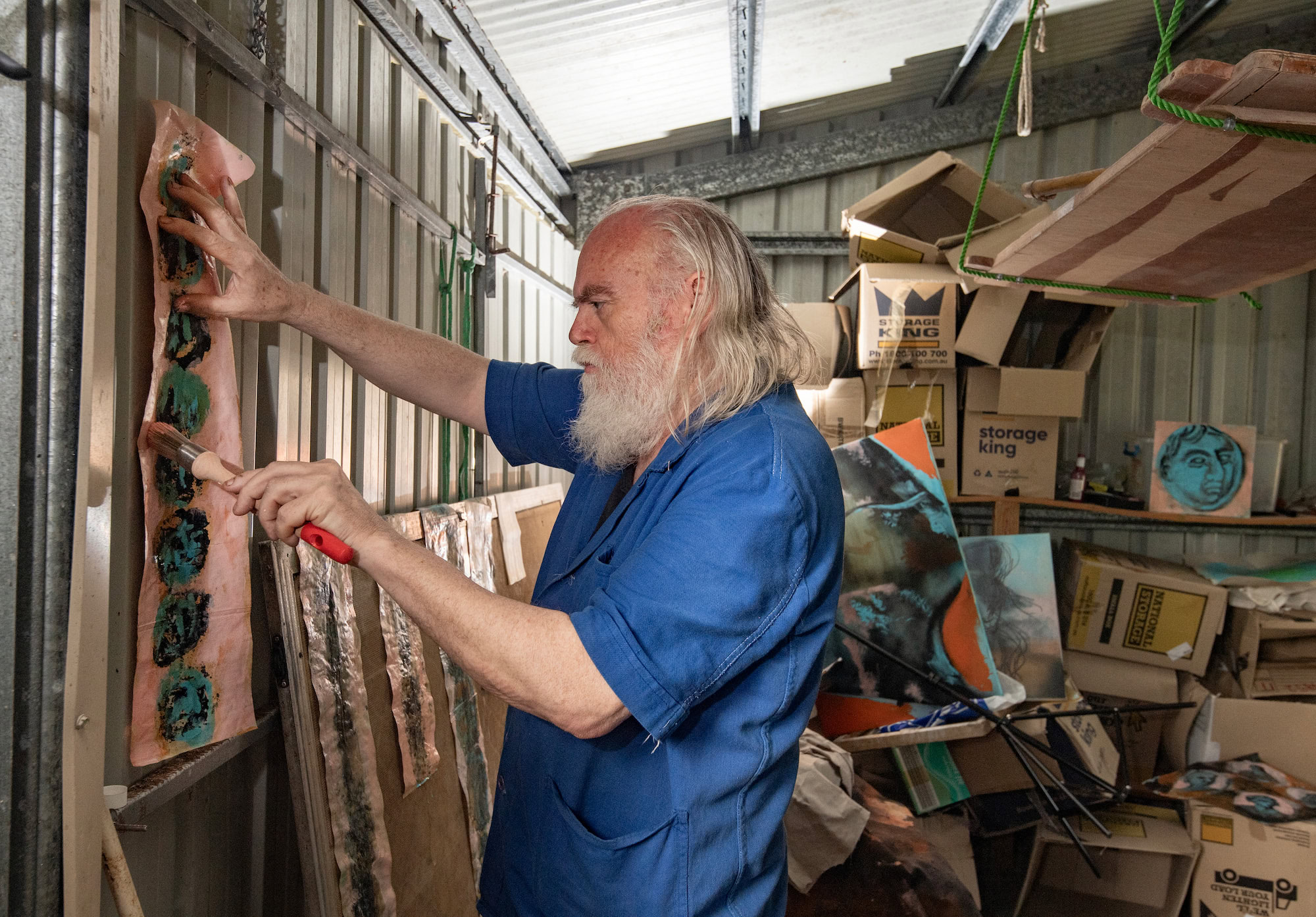 A person with long white hair and beard in a blue shirt applying paint to an artwork in a studio shed