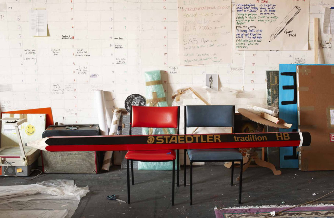 A huge replica of a standard STAEDTLER HB pencil lays horizontally across two chairs. The room surrounding the chairs looks like an artist's studio with materials and objects piled along the back wall, that is also covered with large-scale handwritten notes, including a drawing of a large pencil.