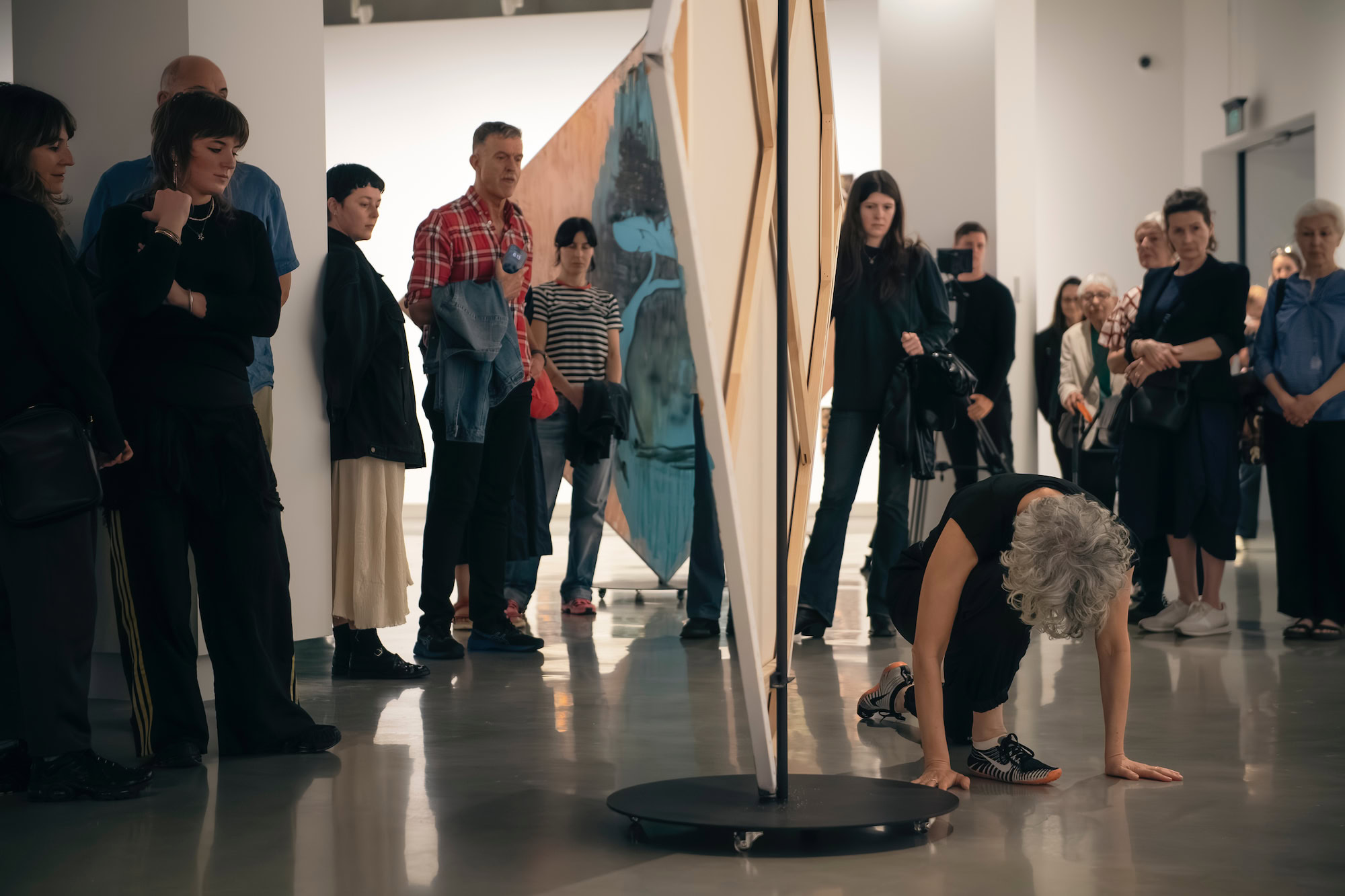 A dancer crouched on one knee next to a painting held up by a stand. Surrounded by people watching them.