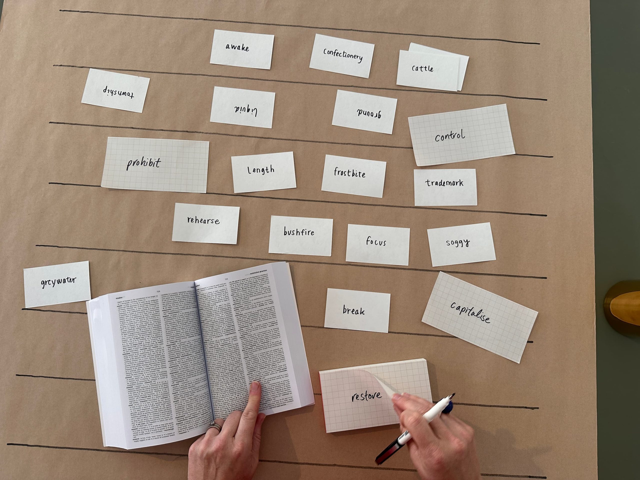 A person holds an open book and a pen with words written on pieces of paper on a table