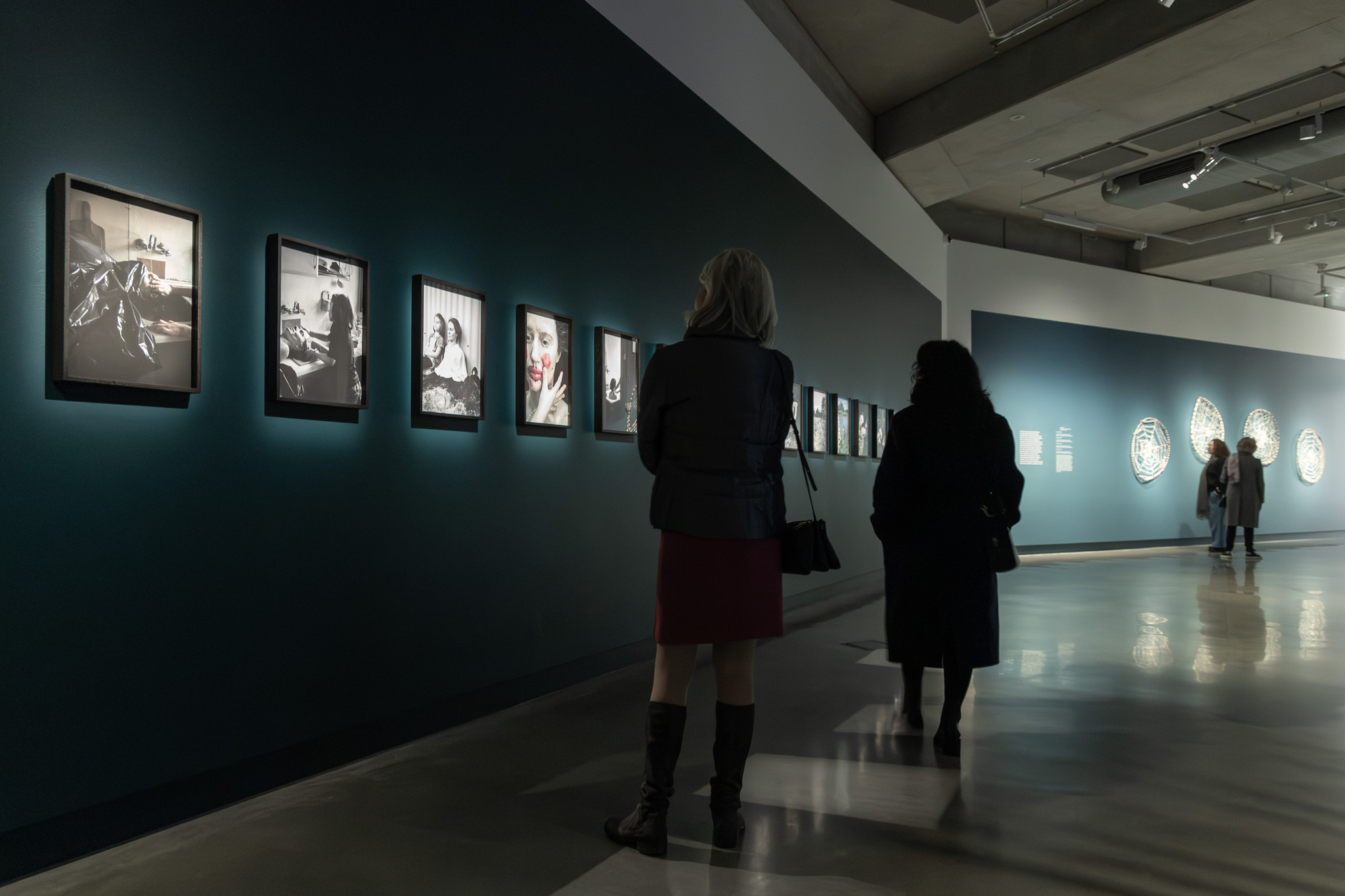 A photograph of a darkened gallery space with two people looking at a row of photographic works