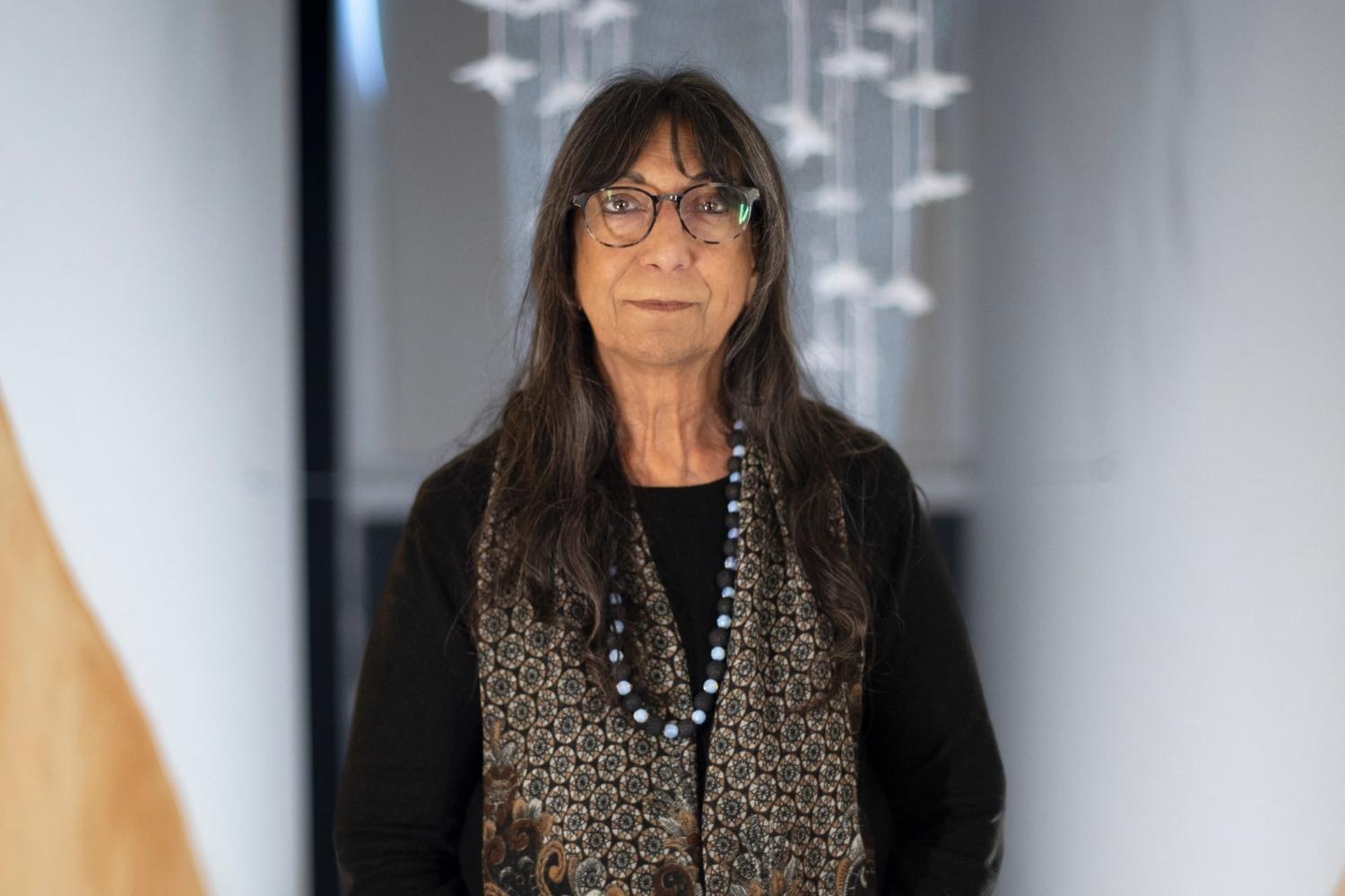 Glenda_Nicholls A photograph of a person with long dark hair wearing dark clothing in front of an artwork featuring suspended feather flowers
