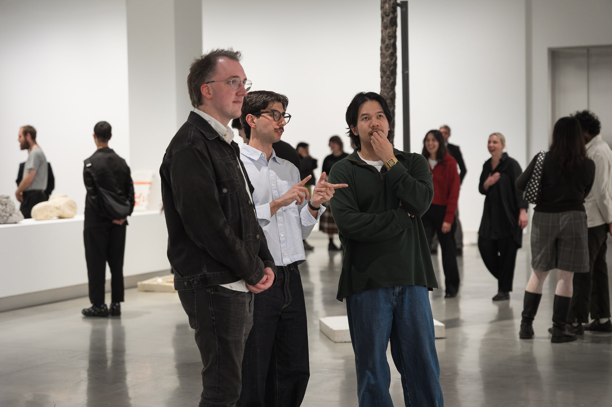 Three people talk in a gallery with other people in background