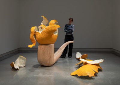 A person walks around a sculpture of an large open orange sitting on a wooden pipe with orange peel on the ground